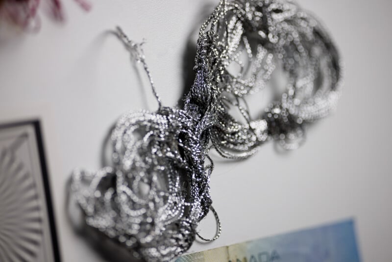 A close-up of tangled silver metallic threads next to the corner of two pieces of paper, one appearing to be an illustration and the other with a printed design.