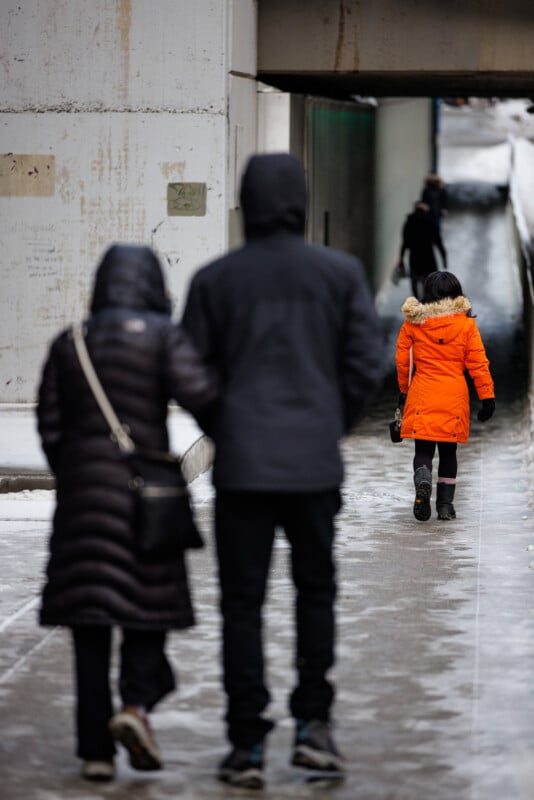 People walk on an icy sidewalk. A child in a bright orange coat walks ahead near a tunnel. Two adults, dressed in dark winter wear, follow behind. Snow and ice are visible on the ground and the surroundings.