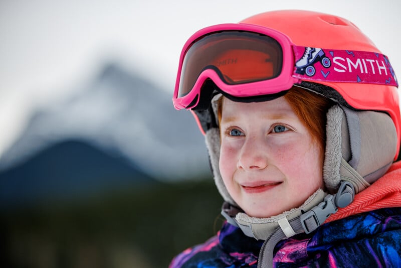 A young child wearing a red ski helmet and goggles smiles warmly. They have red hair and rosy cheeks. In the background, there is a blurred view of a snowy mountain.