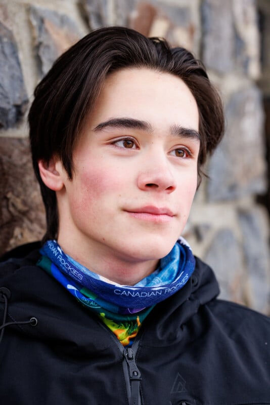 A person with medium-length dark hair is wearing a black jacket and a multicolored neck gaiter with "Canadian Rockies" written on it. They are standing against a stone wall, looking into the distance with a calm expression.