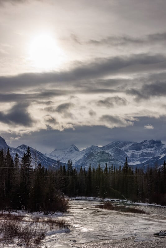 A serene winter landscape with a partially frozen river flowing through a snow-covered forest. Majestic mountains rise in the background under a cloudy sky with the sun peeking through, casting a soft light on the scene.