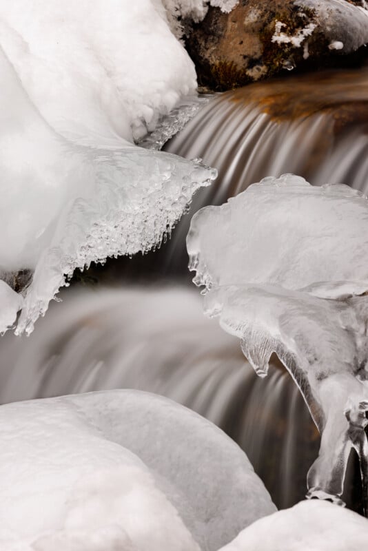 A close-up of icy formations surrounding a small, flowing waterfall. The ice glistens and the water cascades smoothly underneath, creating a dynamic contrast between the solid and liquid elements in a winter landscape.