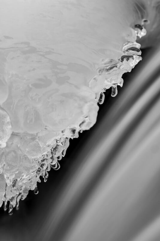 Close-up of icicles hanging from a ledge, with smooth, flowing water in the background. The image is in black and white, highlighting the contrast between the frozen texture of the ice and the fluid motion of the water.