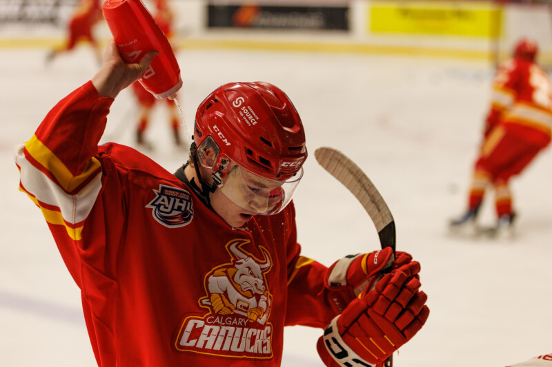 Hockey player in a red Calgary Canucks jersey uses a water bottle, holding it above his head. He wears a helmet with a visor and stands on the ice, with another player visible in the background.