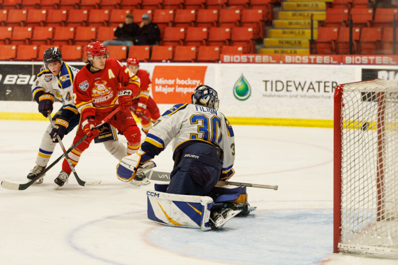 A hockey player in a red uniform attempts to score as an opposing goalie in a white and blue uniform prepares to block the shot. Other players are on the ice, with empty red seats and advertisements in the background.