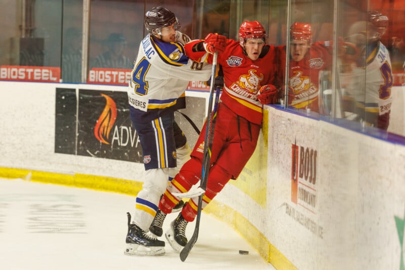 Two hockey players are in action near the rink's glass wall. The player in red is being checked by the player in white and blue. They both hold hockey sticks. The rink has visible advertisements on the boards.