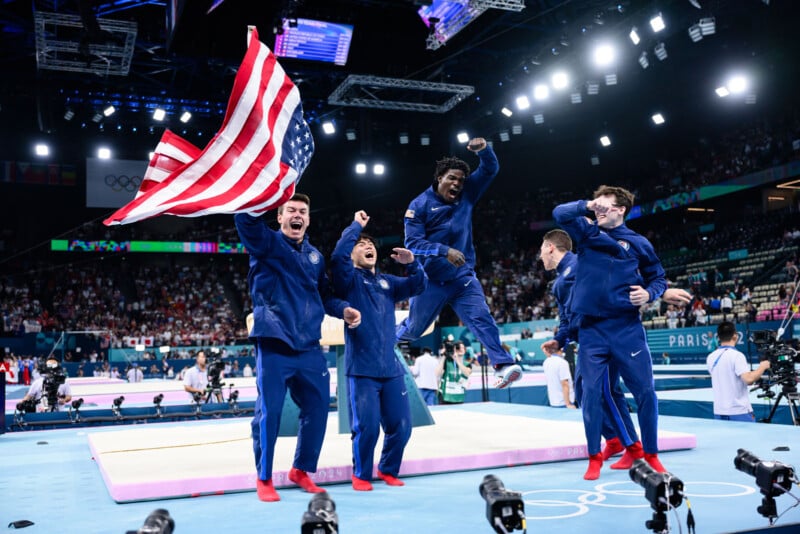 A group of five male athletes dressed in blue Team USA tracksuits celebrate energetically in a gymnastics arena. One athlete holds an American flag. Cameras and audiences surround the scene. The arena is filled with bright lights, spectators, and Olympic branding.