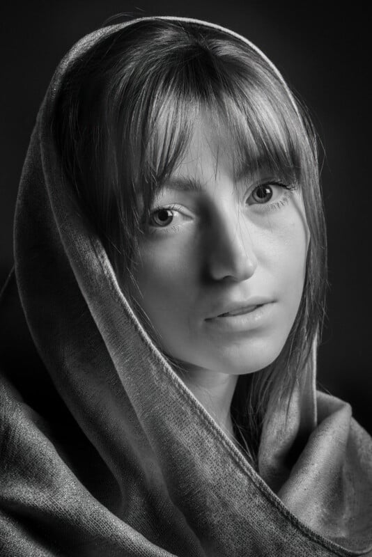 Black and white portrait of a woman with a gentle expression, partially covered by a textured shawl draped over her head. Her eyes look directly into the camera, and the soft lighting highlights her features against a dark background.