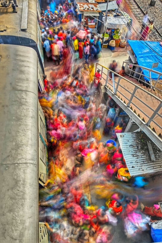 Blurred image of a busy railway station platform with a train on the left. A colorful crowd of people is in motion, creating a vivid swirl of colors. Stalls with goods are set up on the side, and a footbridge can be seen above.