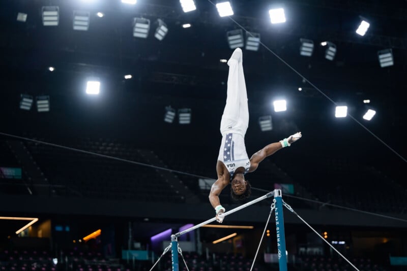 A gymnast performs a handstand on the horizontal bar in a brightly lit indoor arena. The athlete wears a white uniform with blue accents. The background is dark with scattered lights, highlighting the gymnast's focus and strength during the routine.