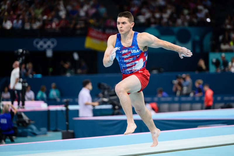 A male gymnast wearing a red, white, and blue USA uniform is mid-routine on the floor exercise at an indoor gymnastics competition. He is performing a dynamic move with one arm forward. The background shows judges and the audience.