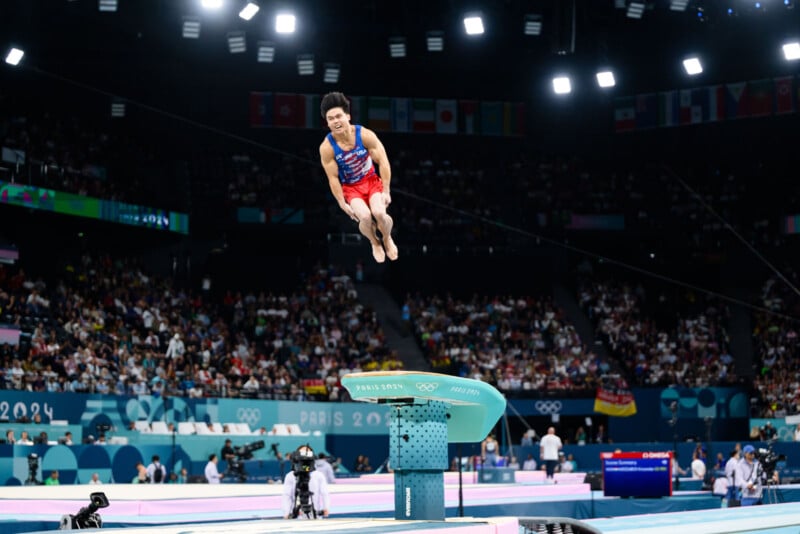 A gymnast performs a mid-air maneuver above the vaulting table at the Olympics, with an audience watching in the background. The arena is brightly lit, and flags from various countries are visible hanging above the audience.