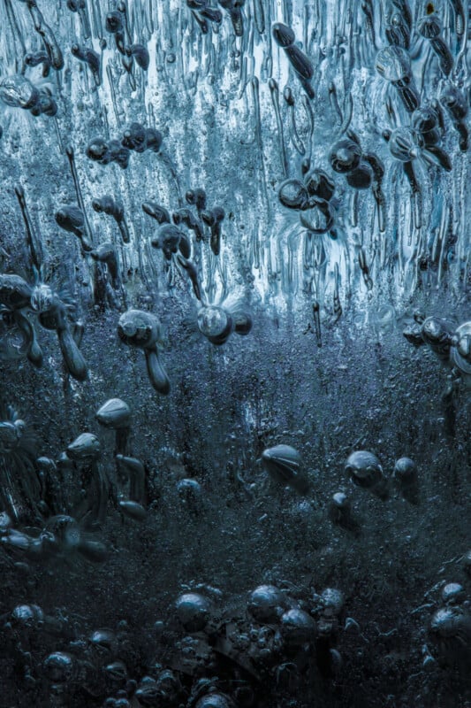 Close-up of ice with trapped air bubbles creating an intricate, abstract pattern. The bubbles vary in size and appear suspended and elongated, giving a sense of motion frozen in time. The overall color is a cool blue tone.