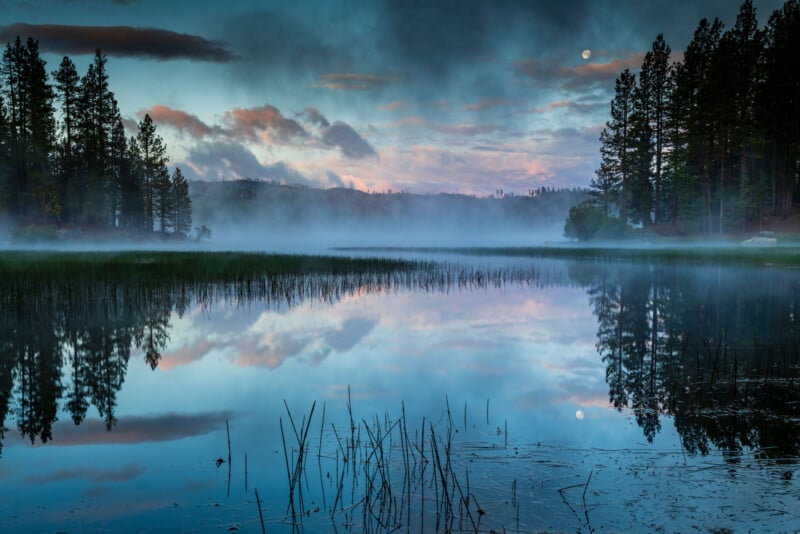 A serene landscape of a misty lake at dawn, surrounded by tall pine trees. The sky has hues of blue and pink, with clouds reflecting on the water. The moon is visible, adding to the calm, scenic atmosphere.