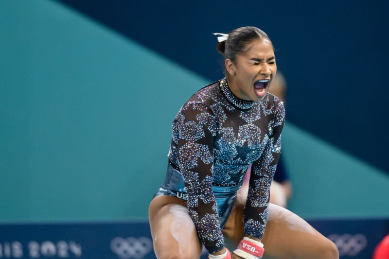 A gymnast in a sparkly, long-sleeved leotard is seen mid-performance with a determined expression. Her hands are wrapped in grips, and she appears to be releasing chalk dust. A blurred background shows part of the gymnasium.