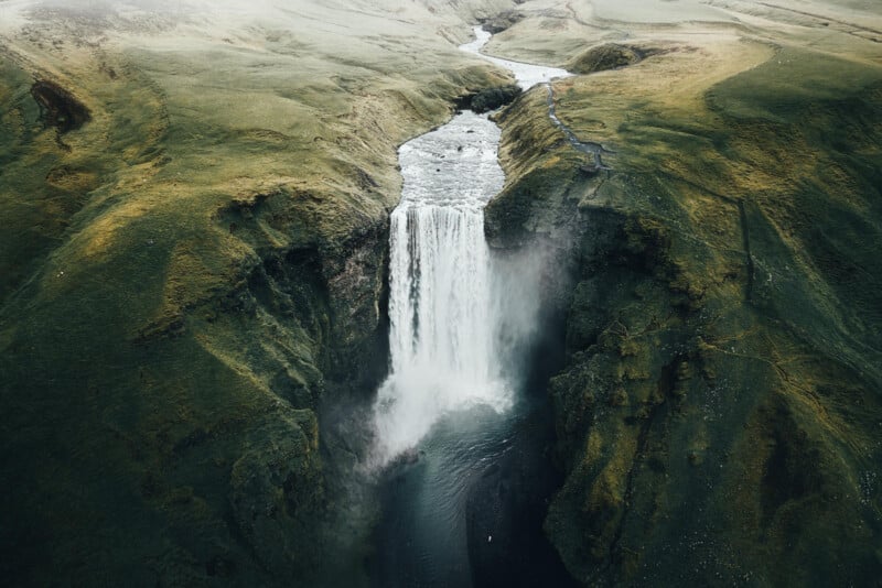 Aerial view of a powerful waterfall cascading between green cliffs into a misty pool below, surrounded by lush, rolling hills. A winding river extends from the waterfall, cutting through the landscape.