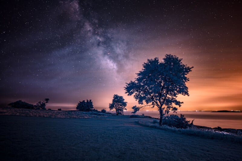 A serene night landscape featuring a starry sky with the Milky Way visible. Silhouetted trees stand near a calm body of water, with a warm orange glow on the horizon, possibly indicating a distant city.