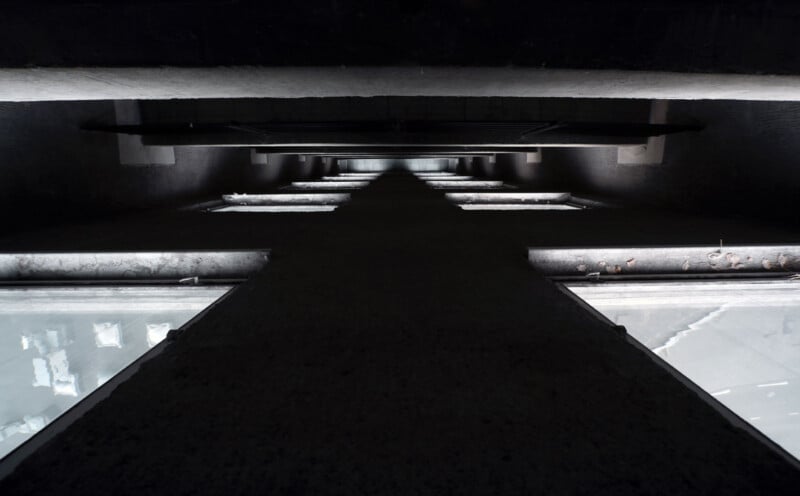 A stark, geometric shadow forms a cross against a dark, textured background. Light filters through rectangular windows on either side, highlighting the shadow's edges and creating a dramatic, symmetrical scene.