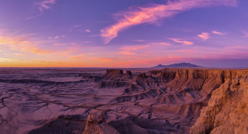 A vast desert landscape at sunset with dramatic rock formations and canyons. The sky is painted in vibrant hues of purple, pink, and orange, casting a warm glow over the rugged terrain and distant mountains.