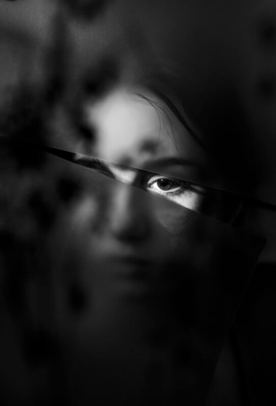 A black and white close-up of a person's face partially obscured by shadows and reflections. Light highlights one eye, creating a mysterious and dramatic effect. The composition includes dark, blurry textures across the image.