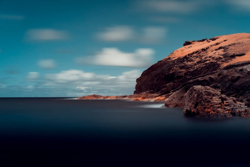 A rocky coastline with textured cliffs under a partly cloudy sky. The water is calm and blends seamlessly with the horizon, creating a serene seascape. The lighting casts a warm hue on the rocks, enhancing the contrast with the blue sky and water.