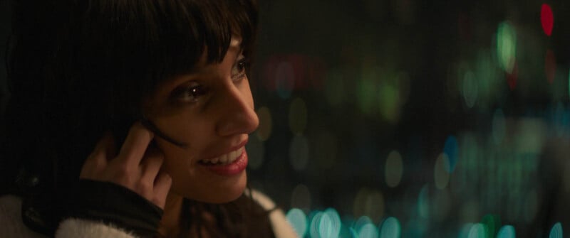 A person with dark hair smiles while holding a phone to their ear. The background is blurred with bokeh lights, suggesting a nighttime urban setting. The person's expression appears happy and engaged in the conversation.