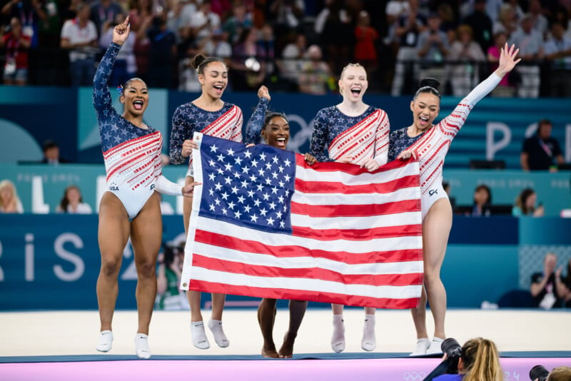 Five female gymnasts in matching patriotic leotards stand together holding an American flag, smiling and celebrating on the podium. The background features a cheering crowd and event signage, indicating a gymnastics competition.