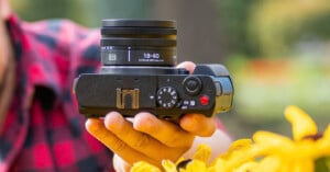 Person holding a black camera with an 18-40mm lens, surrounded by yellow flowers in the foreground. The person is wearing a red plaid shirt, and a blurred outdoor background is visible.