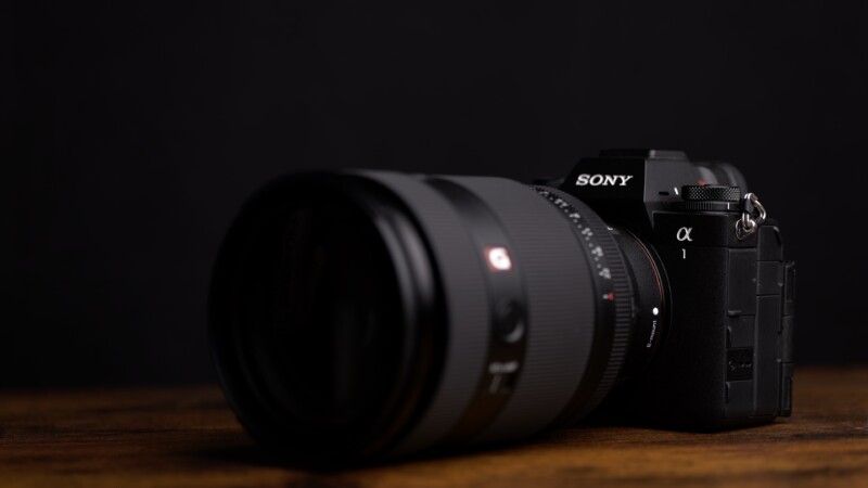 A Sony Alpha mirrorless camera with a large lens is placed on a wooden surface against a dark background. The camera is angled slightly to the left, showcasing its branding and buttons.