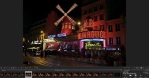 A night view of the Moulin Rouge in Paris, featuring its iconic red windmill and bright neon lights. The building is illuminated against the dark sky, with pedestrians and cars visible in the foreground.