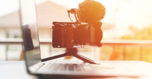 A professional camera with a microphone mounted on a tripod stands in front of a laptop, capturing the screen. The setting is softly lit, creating a warm, sunlit ambiance.