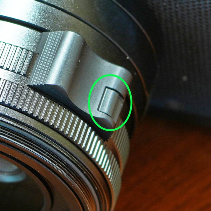 Close-up of a camera lens with a green circle highlighting a small switch on the lens barrel. The lens has a metallic finish and intricate detailing.