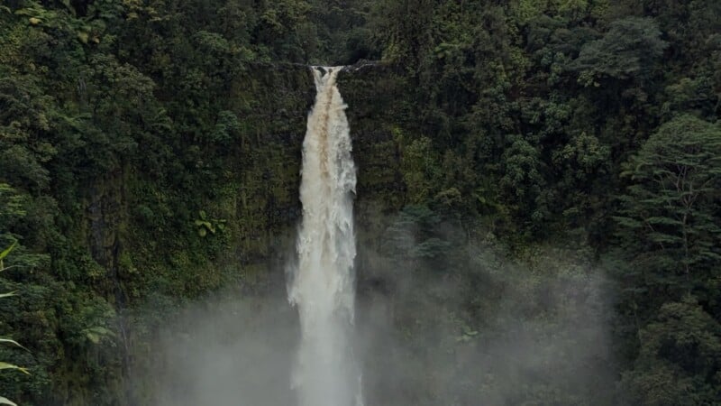 A tall waterfall cascades down a lush green cliff surrounded by dense forest. Mist rises from where the water hits the pool below, blending with the greenery.