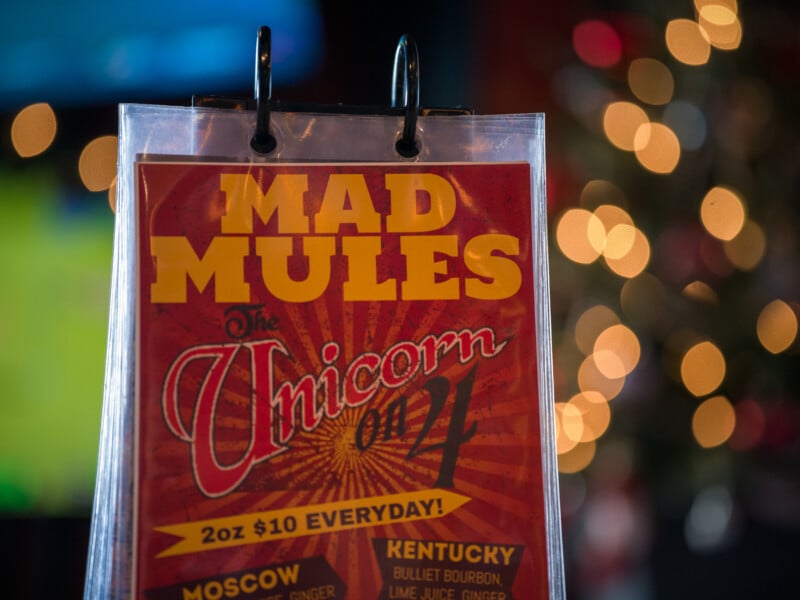 A menu titled "Mad Mules the Unicorn on 7," with drink specials like Moscow and Kentucky, priced at 2oz for $10.00. The background is blurred, featuring round, warm lights.