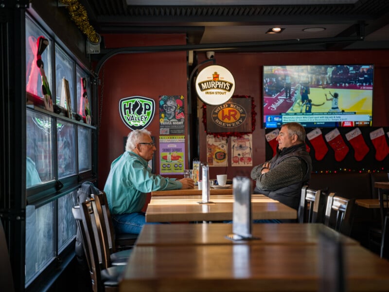 Two men sit across from each other at a wooden table in a cozy pub. The walls are adorned with beer signs, and a TV displays a sports game. Stockings hang along the wall under festive decorations, creating a warm, inviting atmosphere.