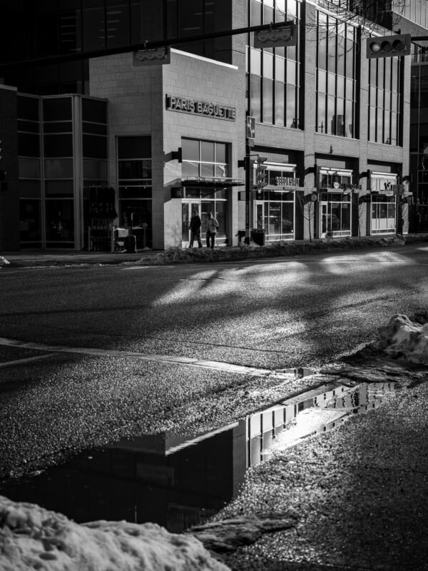 A black and white photo of a city street corner with a "Paris Baguette" storefront. The street is wet, reflecting light, and there's a puddle in the foreground. Two people walk near the store.