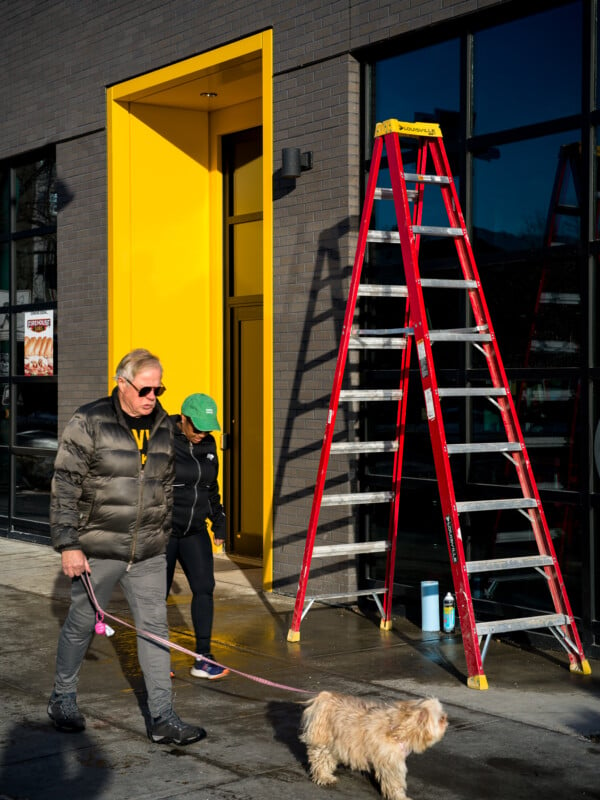 A man in a gray jacket walks a small dog on a leash past a red ladder leaning against a building with a bright yellow doorway. Another person in black workout clothes stands near the entrance.