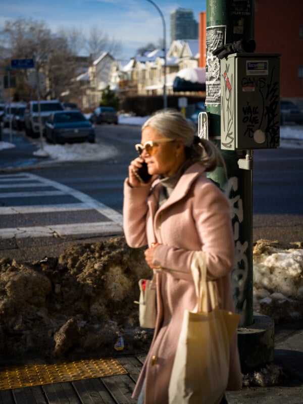 A woman in a pink coat and sunglasses walks down a city street, talking on her phone. She carries a tote bag and is passing by a graffiti-covered utility box. Snow is piled along the curb, and houses are visible in the background.