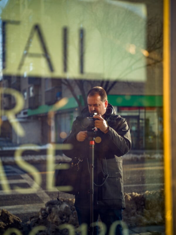 A person taking a photo through a glass window with a camera on a tripod. The reflection shows part of a street scene outside, with buildings and a hint of a snowy landscape. Text on the glass is partially visible.