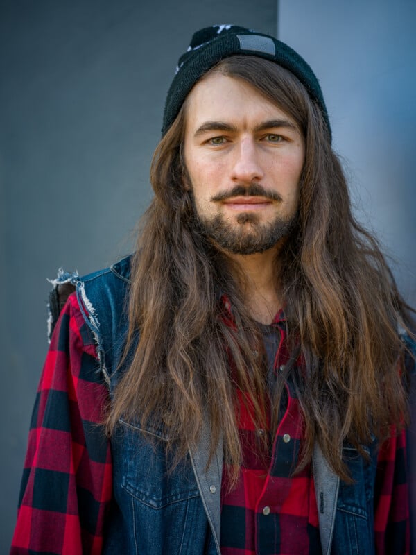 A person with long brown hair and a beard wearing a black beanie, red and black plaid shirt, and a denim vest stands in front of a gray background, looking directly at the camera.