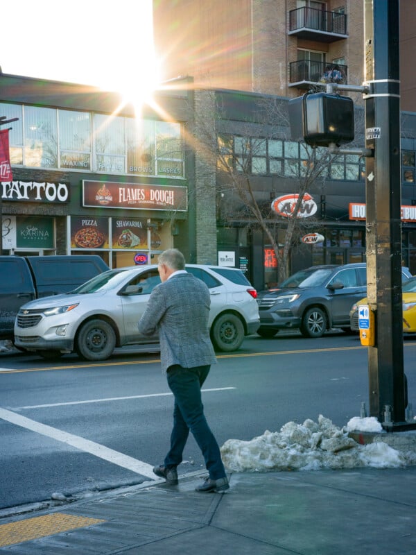 A man in a gray blazer crosses a city street at a crosswalk. Cars are lined up on the road, and a building with storefronts sits in the background. The sun shines brightly, creating a flare effect. Snow is piled along the sidewalk.