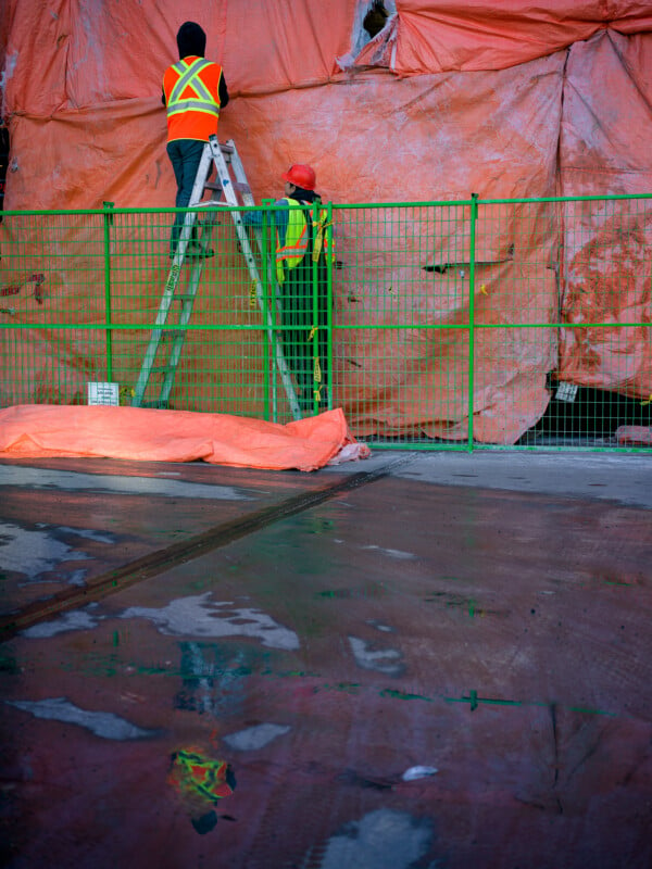 Two construction workers in safety gear stand on a ladder against an orange tarp-covered fence. One worker holds the ladder while the other works above. A puddle on the ground reflects their image and the bright orange covering.