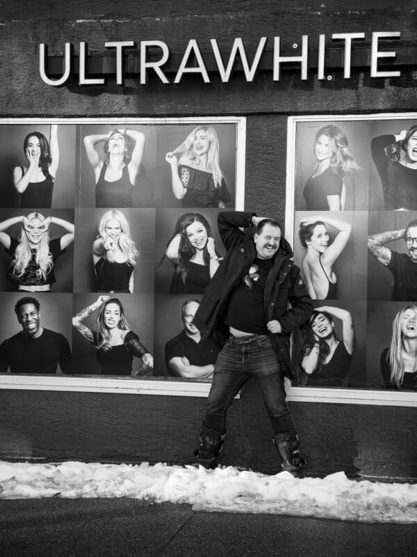 A person poses playfully in front of a billboard displaying black-and-white portraits of various people. Snow is visible on the ground, and the billboard has "ULTRAWHITE" written at the top.