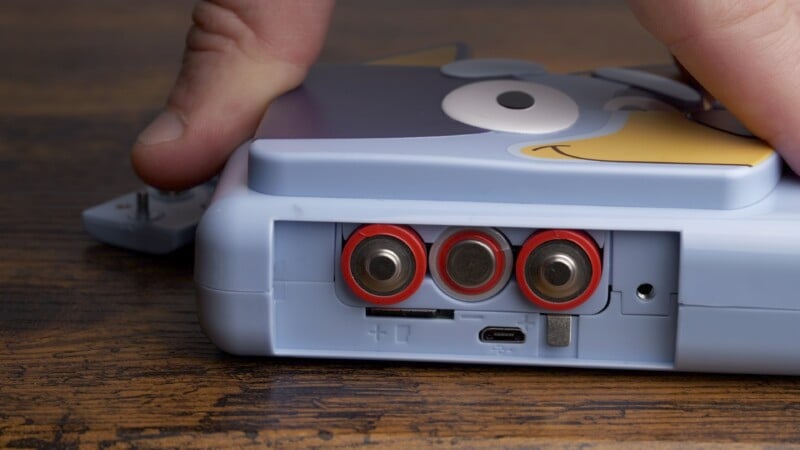 A person is inserting batteries into a blue electronic device with cartoon eyes on it. The focus is on the battery compartment, which has three batteries aligned horizontally. The device is placed on a wooden surface.