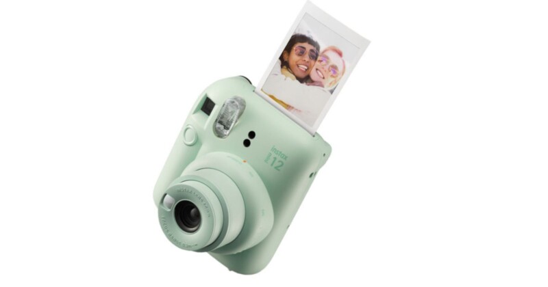 A mint green instant camera with a printed photo emerging from the top. The camera features a lens at the front and a small flash. The printed photo shows two smiling people.