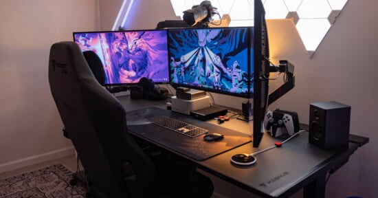 A modern gaming setup featuring two large monitors displaying vibrant graphics, a gaming chair, and various peripherals including a keyboard, mouse, speakers, headset, and a gaming controller. The setup is illuminated with ambient lighting.