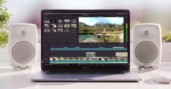 A MacBook Pro with video editing software open on the screen, showing a timeline and a preview of a person sitting on a pier by a lake with mountains in the background. Two white speakers flank the laptop on a white desk near a window with greenery outside.