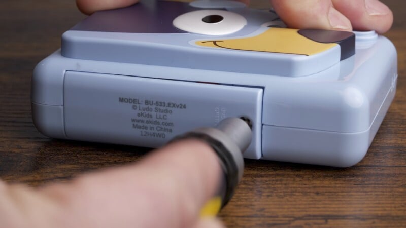 Close-up of a person using a screwdriver to open the battery compartment of a blue electronic device with a cartoon face on it. The device is on a wooden surface, and the text on the back indicates the model number and manufacturer.