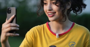 A smiling person with curly hair taking a selfie outdoors, wearing a yellow shirt with a red collar and a small pin microphone clipped to the shirt.