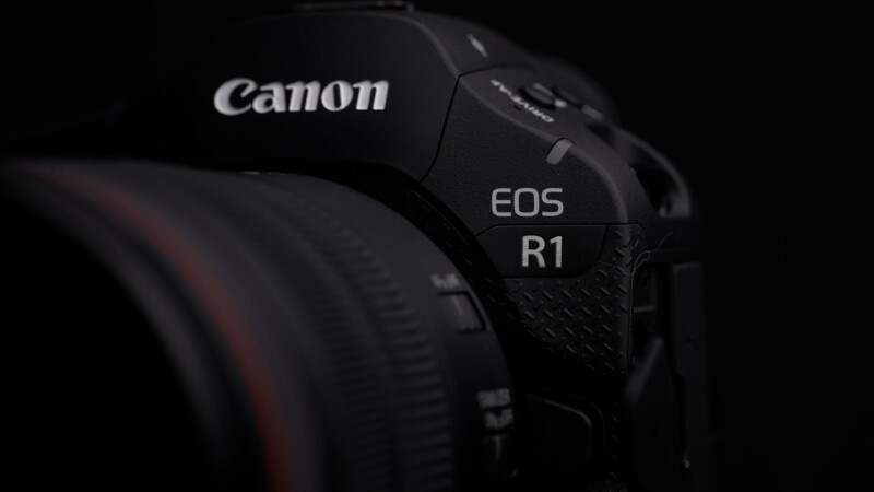 Close-up of a Canon EOS R1 camera, focusing on the camera's branding and body details. The image is predominantly dark, highlighting the camera's sleek design and lettering on the upper right side.